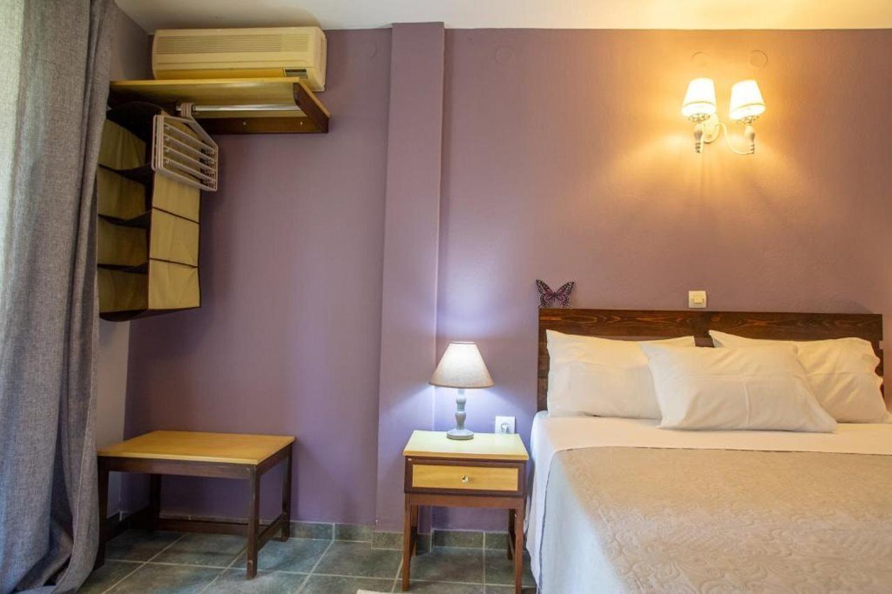 A hotel room with a bed, a nightstand with a lamp, a small table, a shelf with hanging clothes, and a wall-mounted air conditioner.