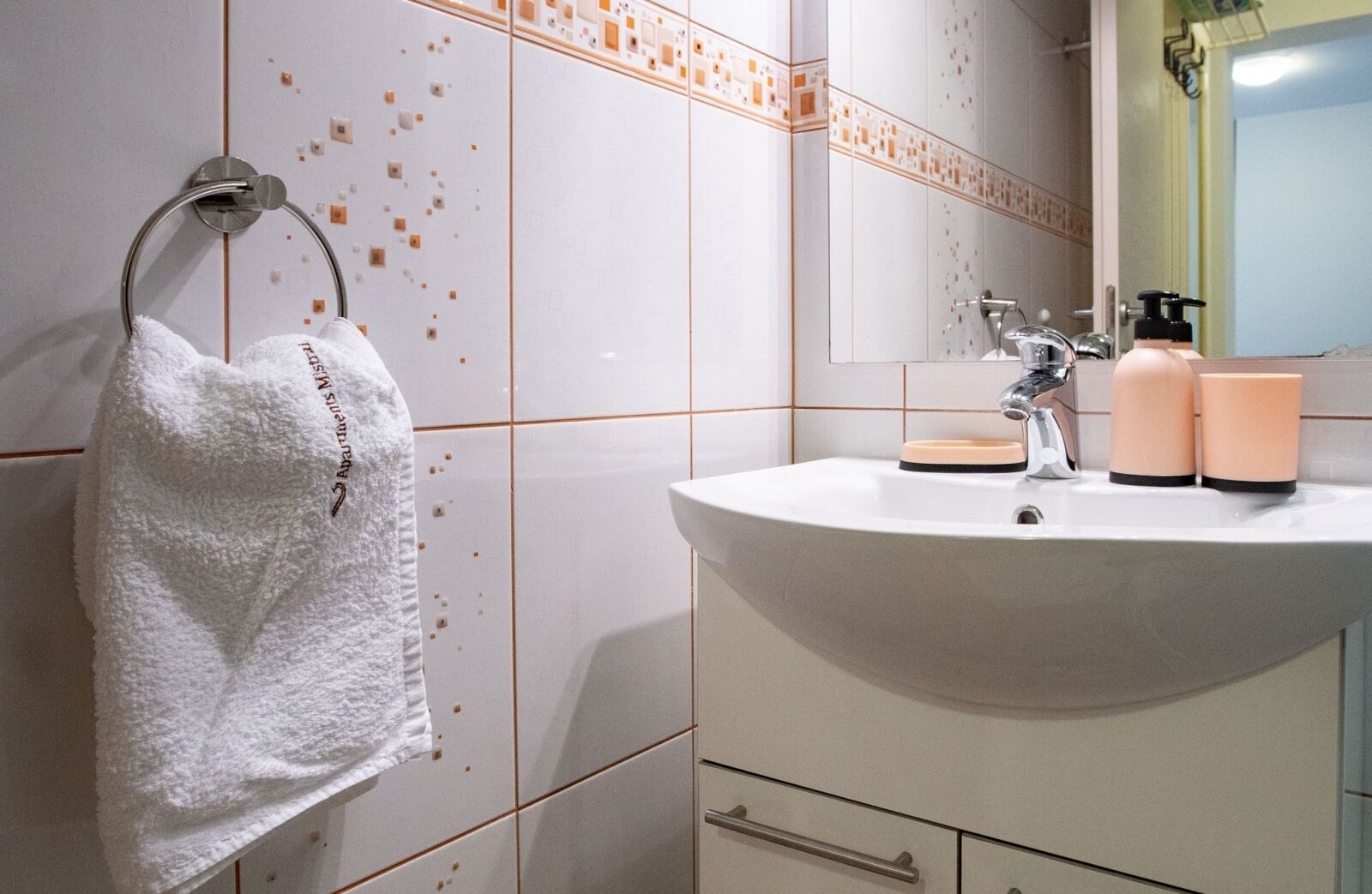 Un baño con un lavamanos blanco, una toalla colgada en un aro de metal y un espejo sobre el lavamanos.