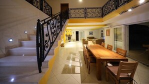 A spacious dining area with a long wooden table and chairs, a staircase with ornate black railings, and a bright, well-lit interior.