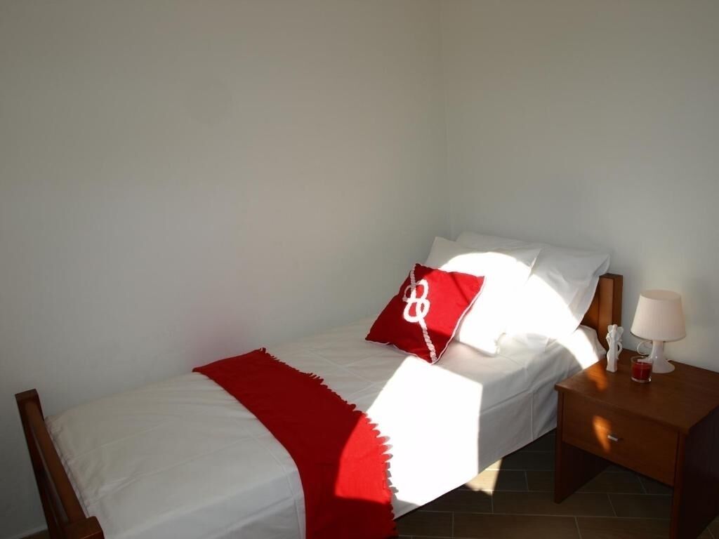 A single bed with white bedding and a red throw blanket, a nightstand with a lamp, and a small red object on the bed.