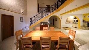 A spacious dining area with a wooden table and chairs, a staircase with black railings, and a kitchen area with yellow walls.