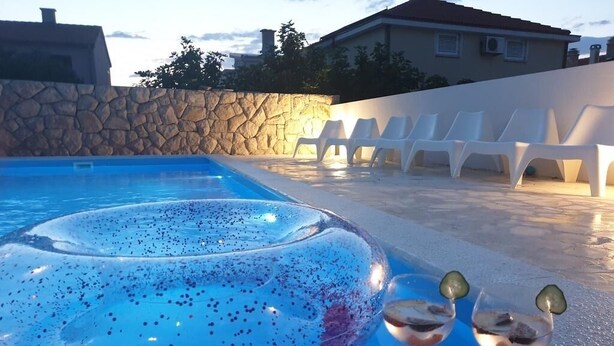 A swimming pool with a hot tub, white lounge chairs, and two glasses of drinks on a textured surface.