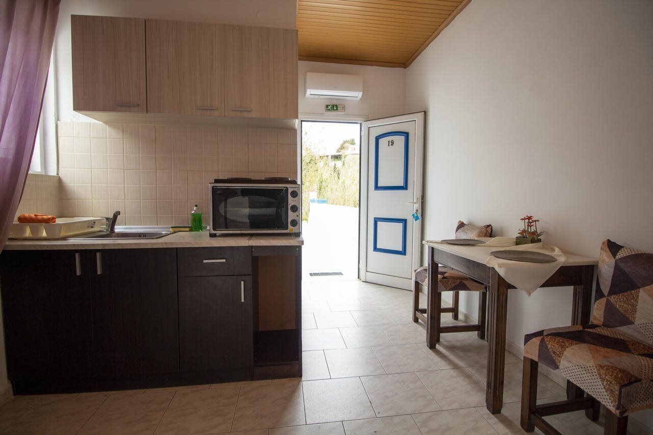 Een keuken met een magnetron, spoelbak en kastjes, een eettafel met stoelen, en een deur met het nummer 19.