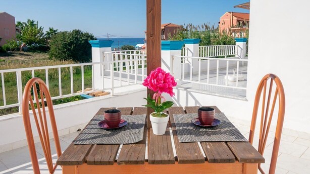 Mesa de madera con dos sillas, una planta en maceta y dos tazas en un balcón con vistas a la costa.