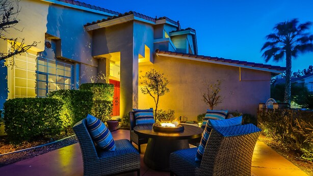 An outdoor patio with wicker furniture and a fire pit, surrounded by a well-maintained garden and a house with a tiled roof.