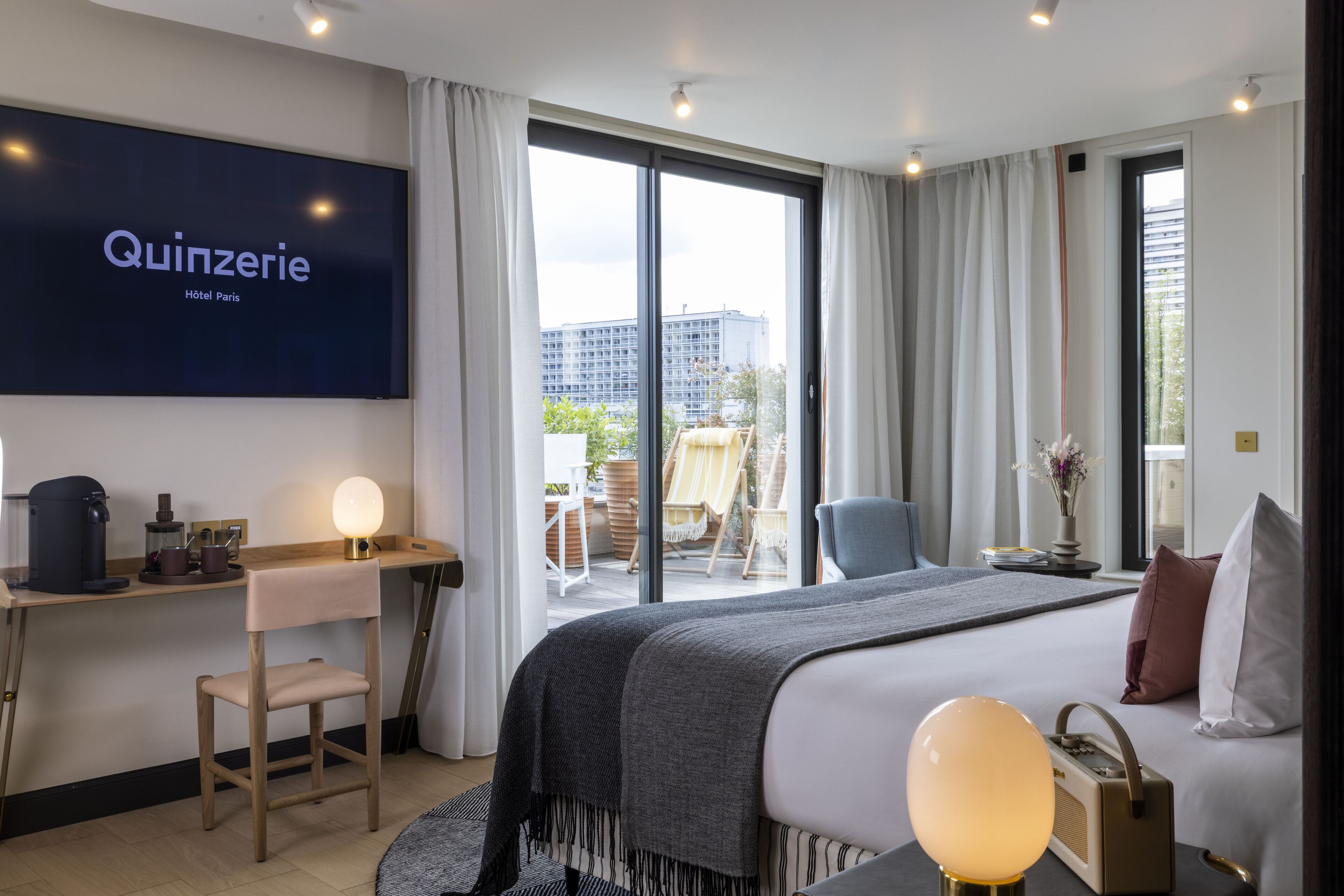 Une chambre d’hôtel équipée d’un lit, d’un bureau, d’une chaise, d’un téléviseur affichant le nom « Quinzerie Hôtel Paris », et d’un balcon avec du mobilier d’extérieur.
