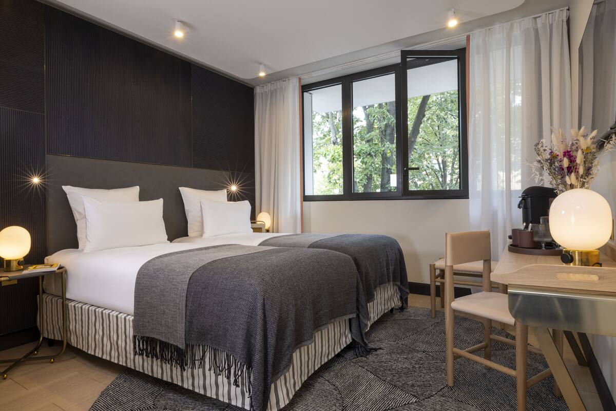 A modern hotel room with a large bed, bedside tables with lamps, a desk with a chair, and a window with a view of trees.