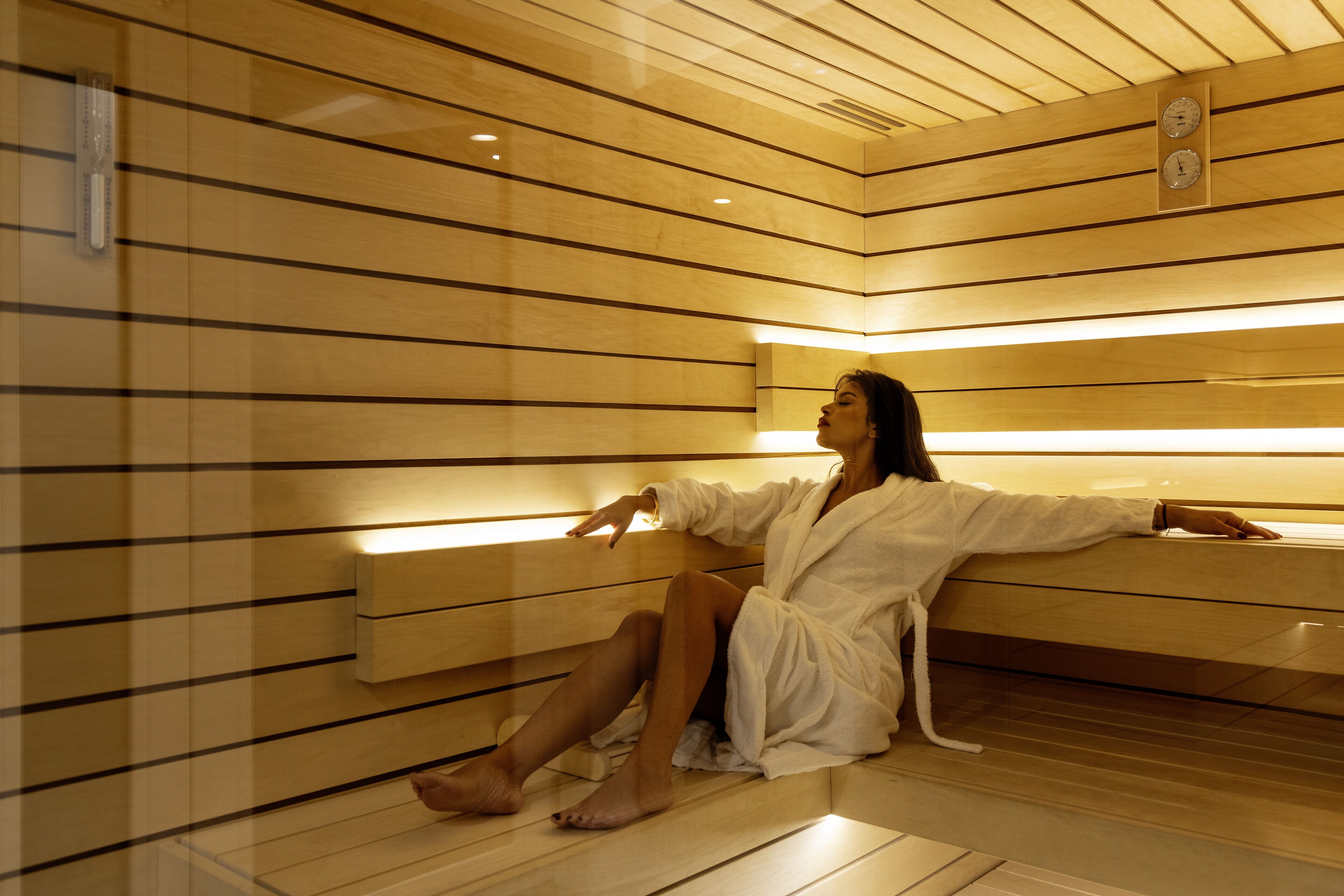 Una persona con bata blanca sentada en un banco de madera dentro de un sauna con paredes y techo de madera.