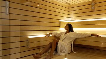 Una persona con bata blanca sentada en un banco de madera dentro de un sauna con paredes y techo de madera.
