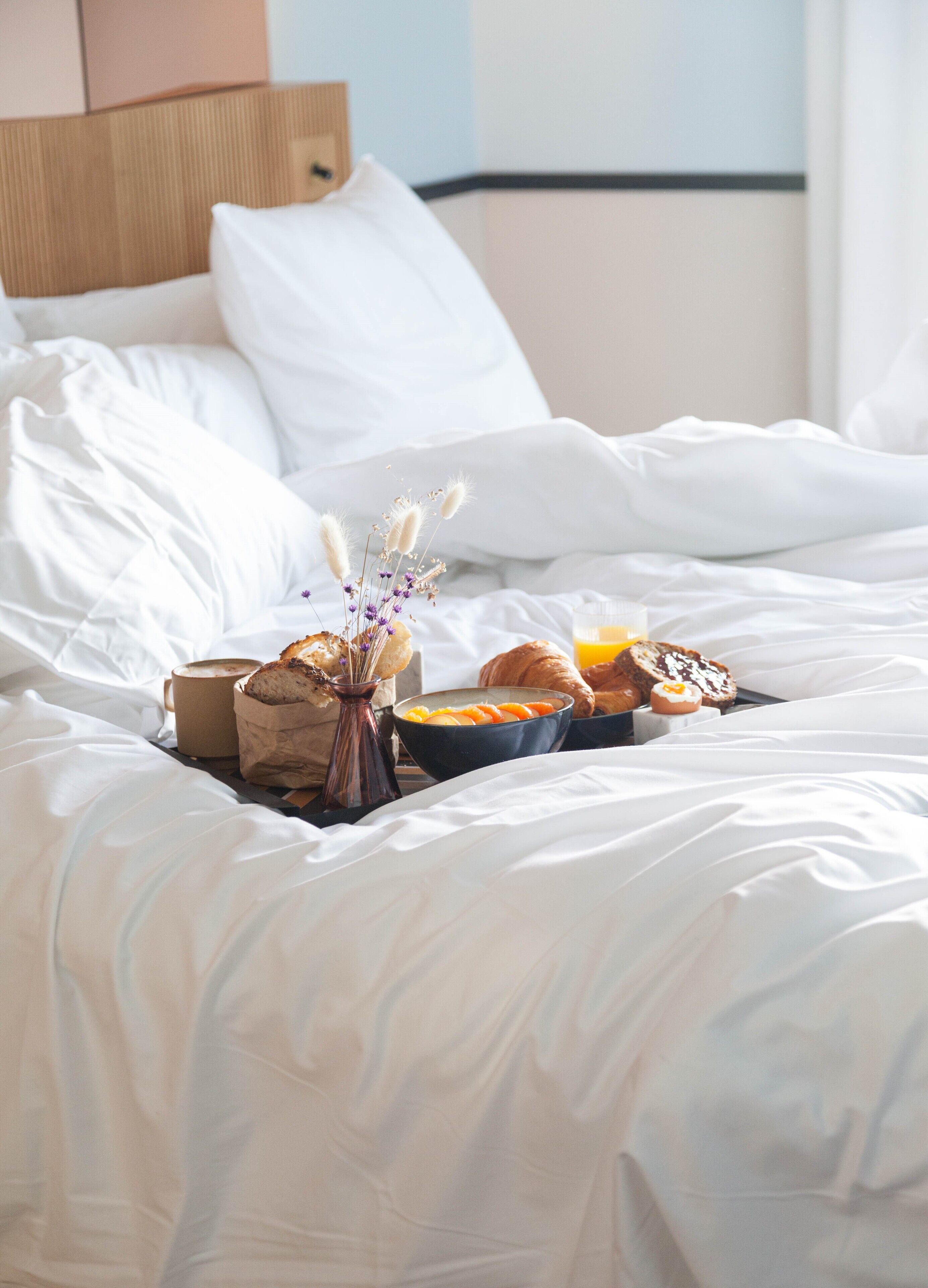 Una cama bien hecha con sábanas blancas, un florero con flores secas, una taza de café, croissants y un bol de fruta.
