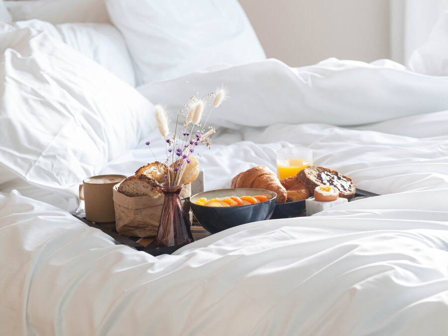 Una cama bien hecha con sábanas blancas, un florero con flores secas, una taza de café, croissants y un bol de fruta.