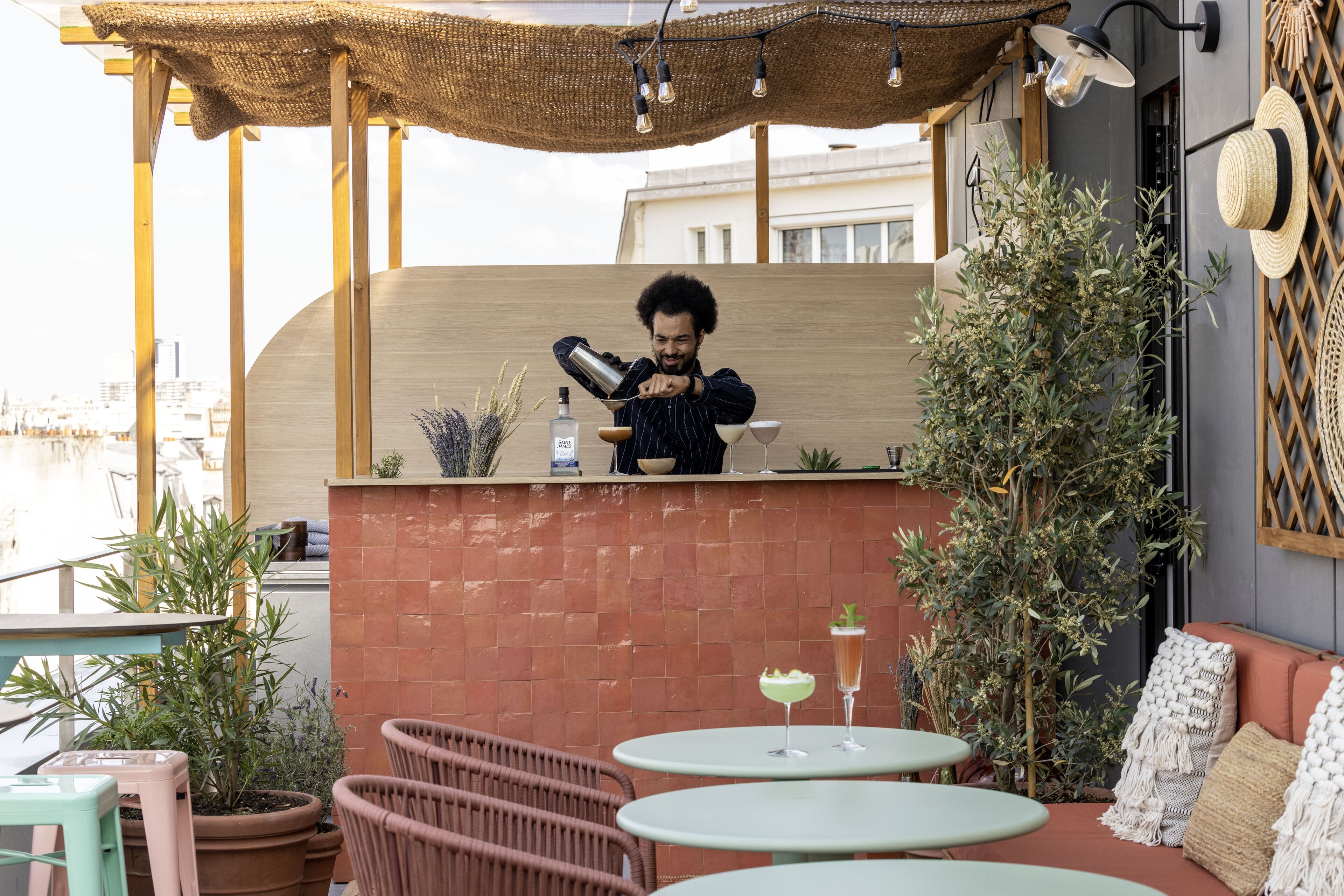 A terrace with round tables, wicker chairs, and a bar area where a person is pouring a drink.