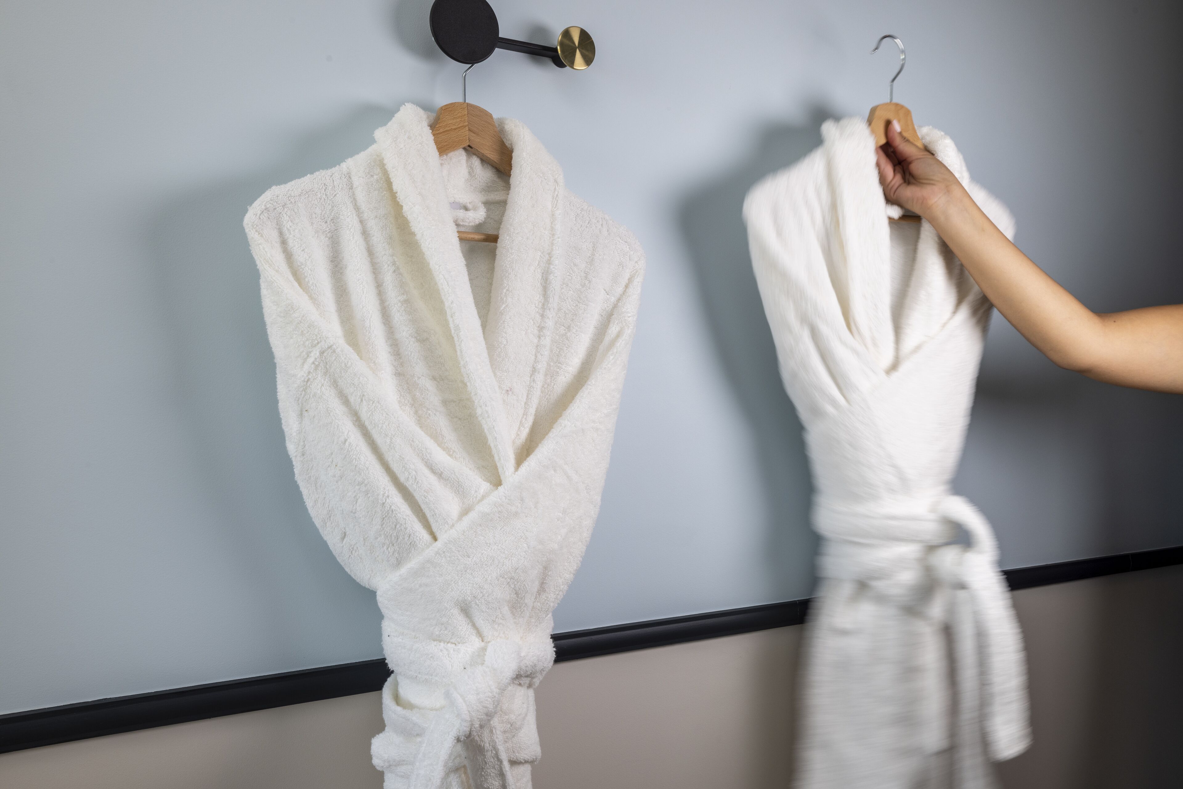 Two white bathrobes hanging on a wall, one on a wooden hanger and the other on a metal hook.