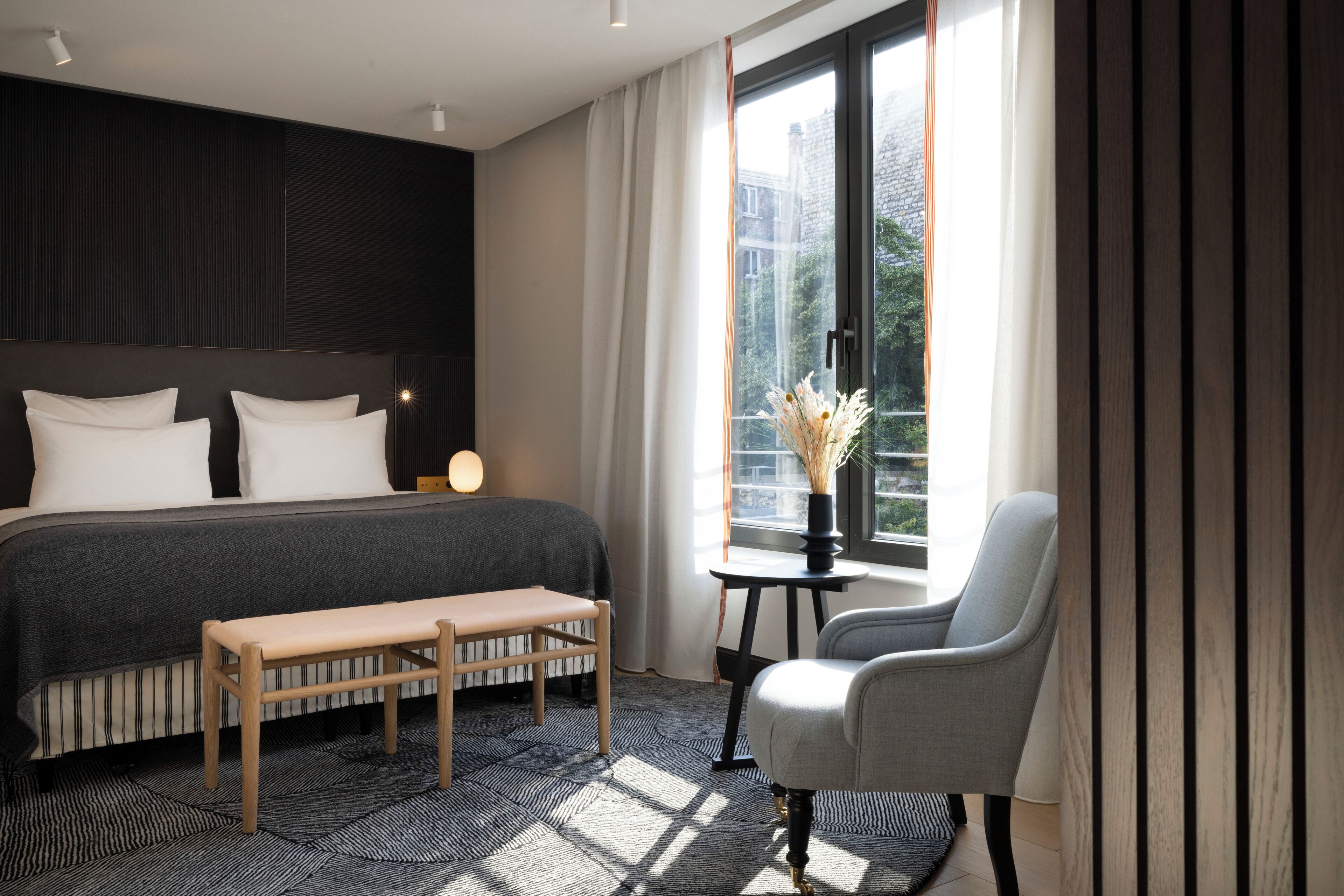 A modern hotel room with a large bed, a bench, a chair, and a view of the city through a window.