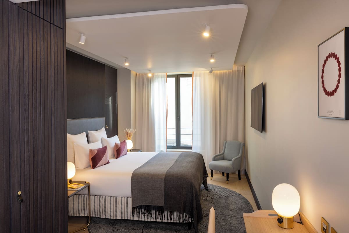 A modern hotel room with a large bed, bedside tables, a chair, a TV, and a view of the outdoors.