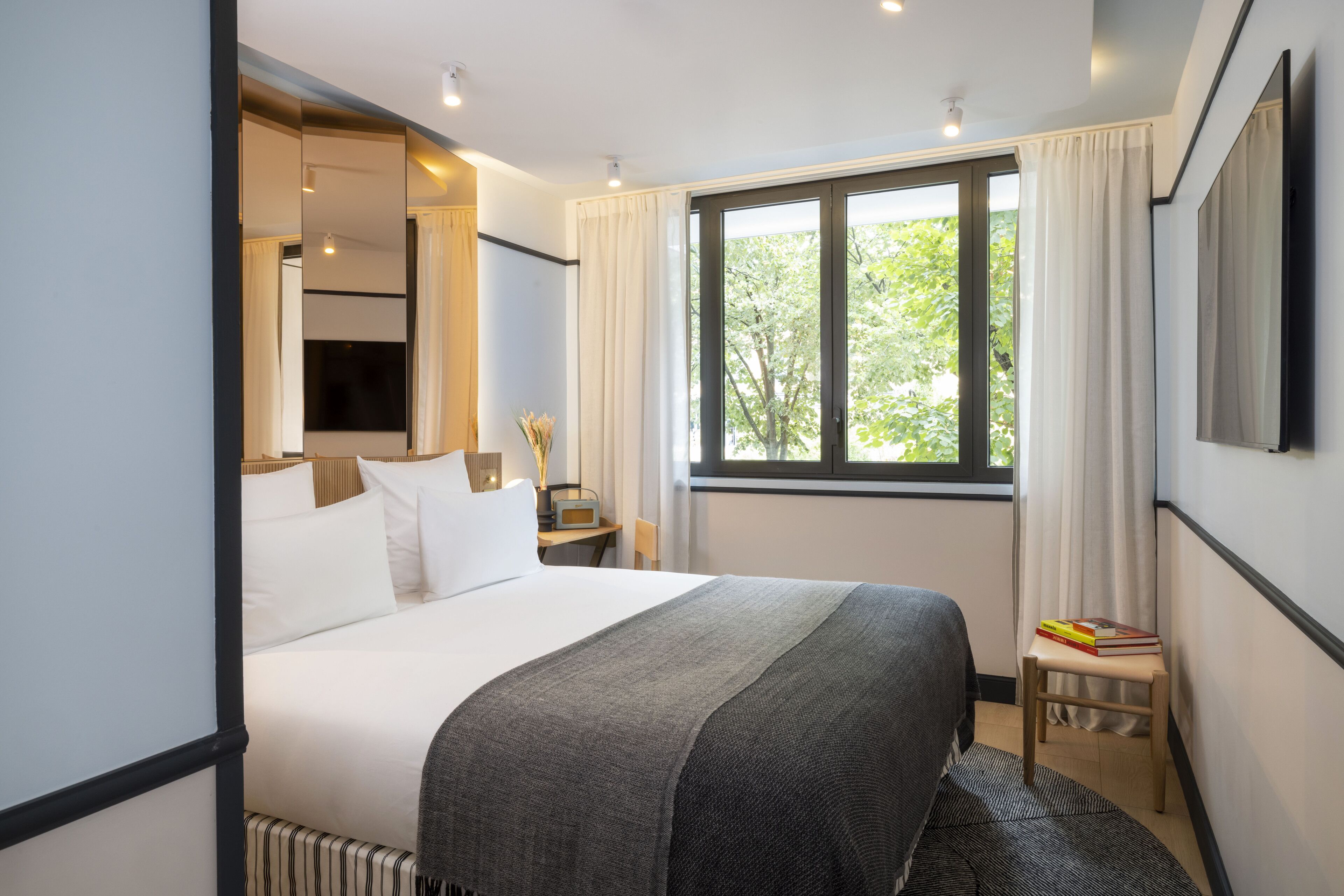 Una habitación de hotel moderna con una cama grande, una mesita de noche con una lámpara, una pequeña mesa con libros y una ventana con cortinas.