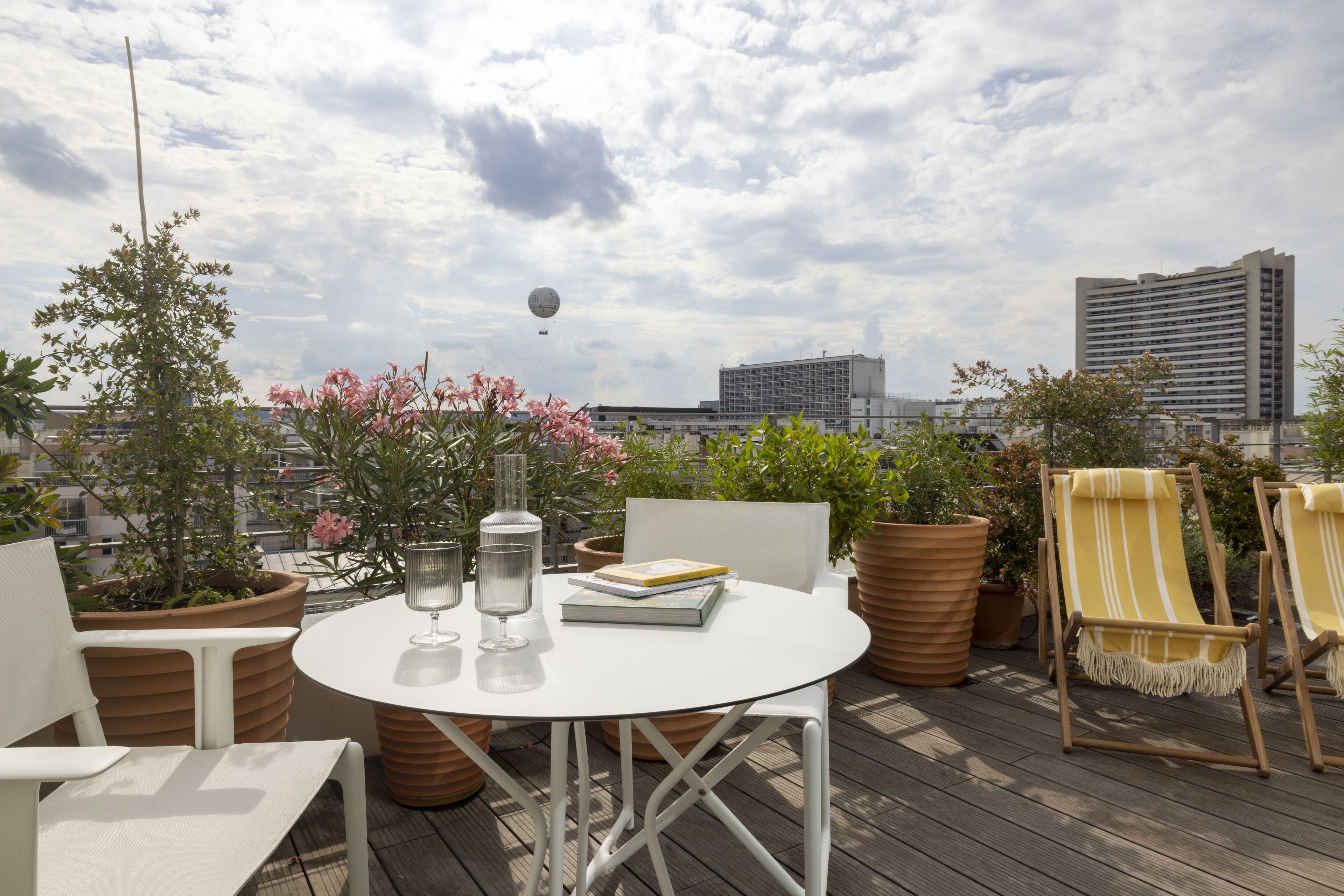 A rooftop terrace with a round table, chairs, potted plants, and a view of the city skyline.