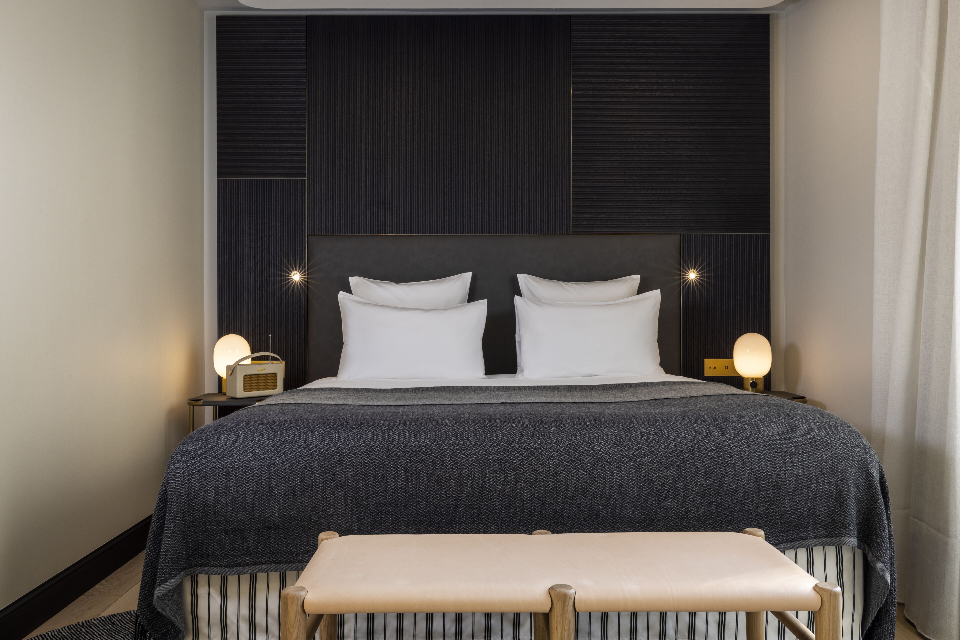 Una habitación de hotel moderna con una cama grande, dos mesitas de noche con lámparas, un banco y un cabecero oscuro.