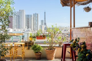 A balcony with potted plants, wicker furniture, and a view of a cityscape including tall buildings and a tower. - Quinzerie Hotel (Paris)