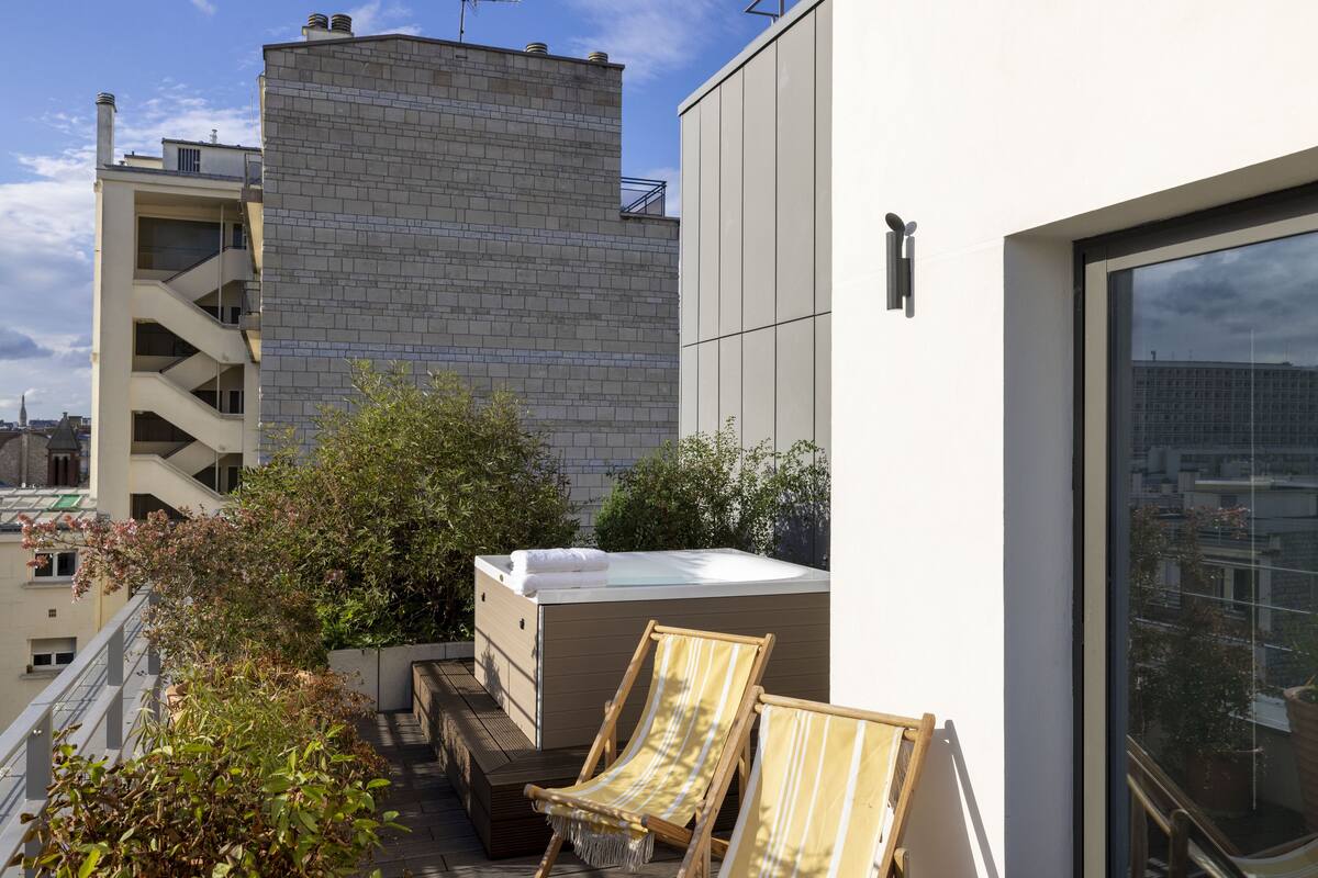 A rooftop terrace with a hot tub, two striped lounge chairs, and a view of the cityscape.