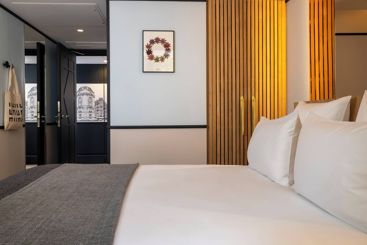 A hotel room with a bed, a framed artwork of a wreath, a wooden slat wall, and a view of a building through a door.