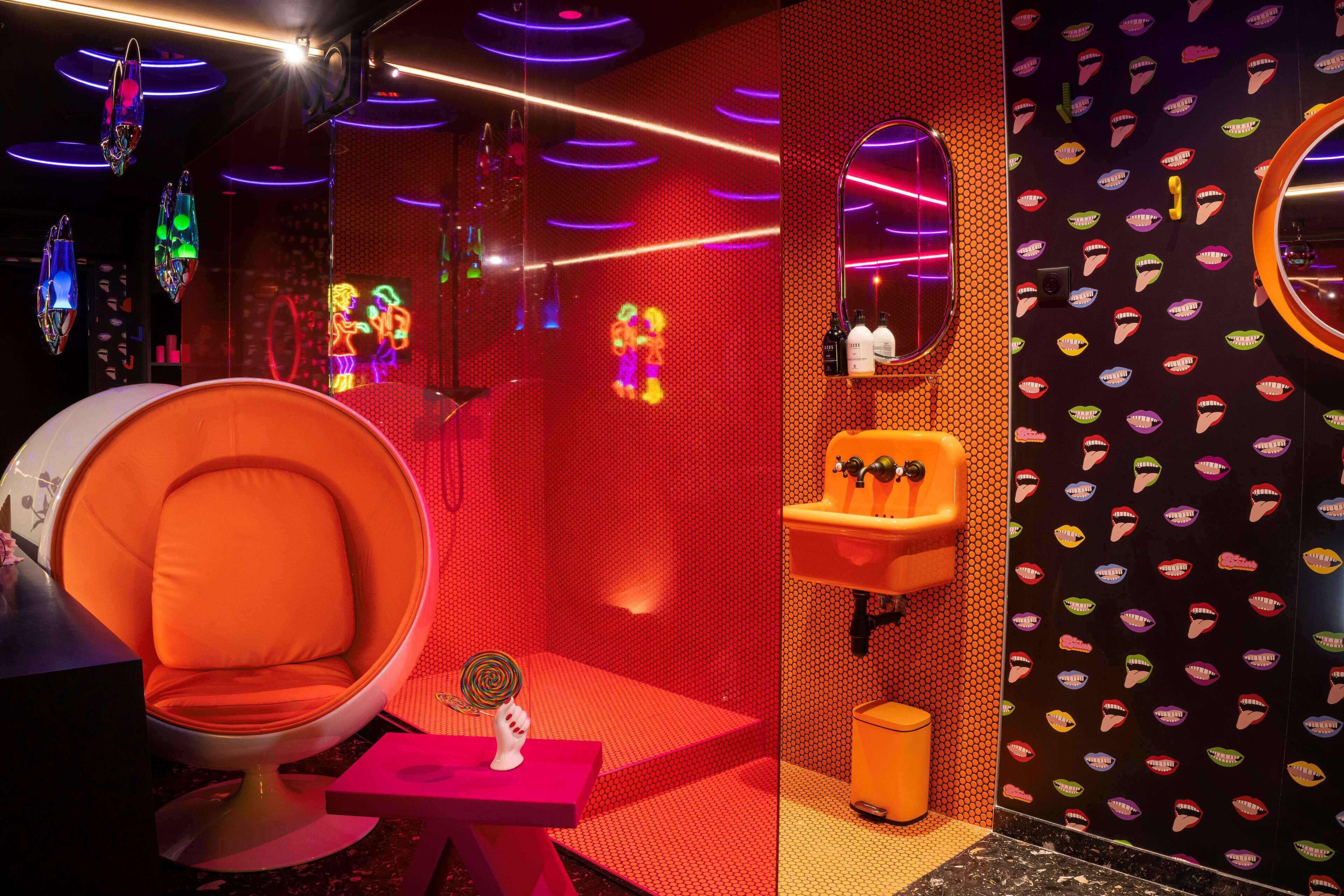 A modern bathroom with a red and orange color scheme, a round orange chair, a small table with a lollipop, a sink, and a mirror with a unique design.