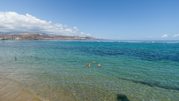 A clear coastal view with calm waters, a sandy beach, and distant hills.