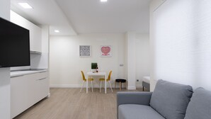 A modern living room with a grey sofa, a dining area with a white table and yellow chairs, and a kitchen with white cabinets and a flat-screen TV.