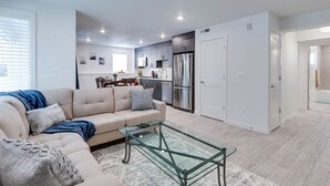 A modern living room with a sofa, a glass coffee table, and a kitchen area with a refrigerator and cabinets.