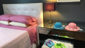 A bedroom with a white tufted headboard, pink patterned bedding, a bedside lamp, and a small stool with flip-flops.