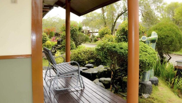 A wooden deck with a metal chair, surrounded by lush greenery and a small pond. - VILLA ADE - 0224 KOTA BUNGA PUNCAK (Cipanas)