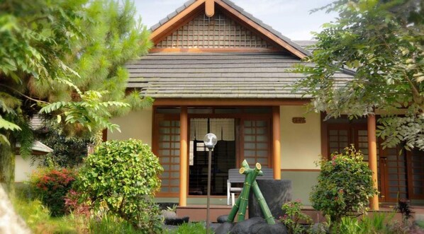 Une maison traditionnelle de style japonais, avec un toit recouvert de tuiles, des poutres en bois et un petit jardin agrémenté de diverses plantes et d’une lanterne en pierre.