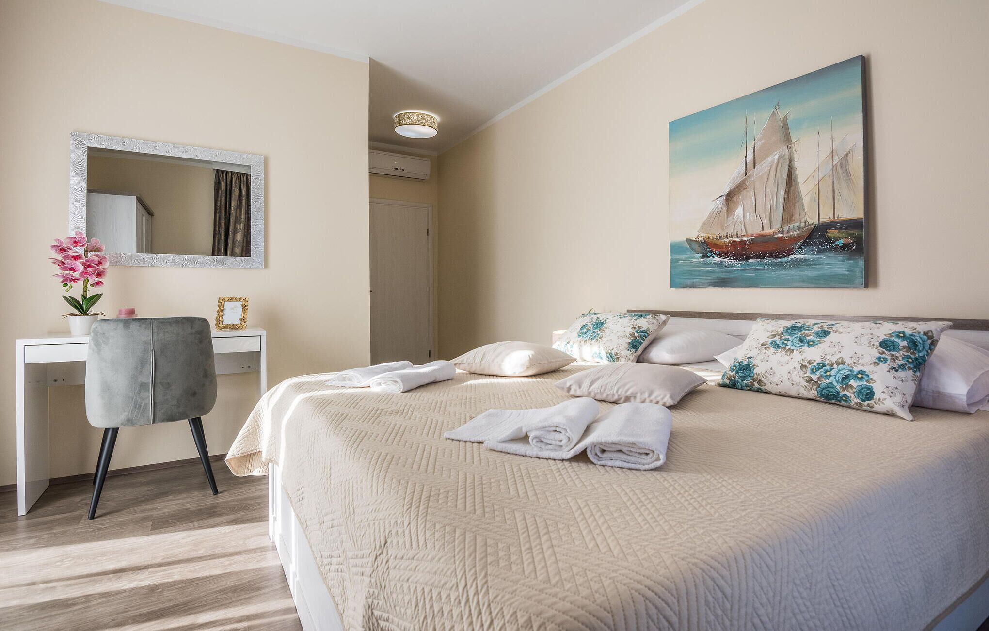 Ett hotellrum med en säng, ett skrivbord, en stol, en spegel och en tavla som föreställer en segelbåt.