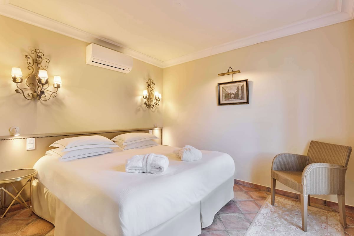 A hotel room with a large bed, two wall sconces, an air conditioner, a framed picture, and a chair.