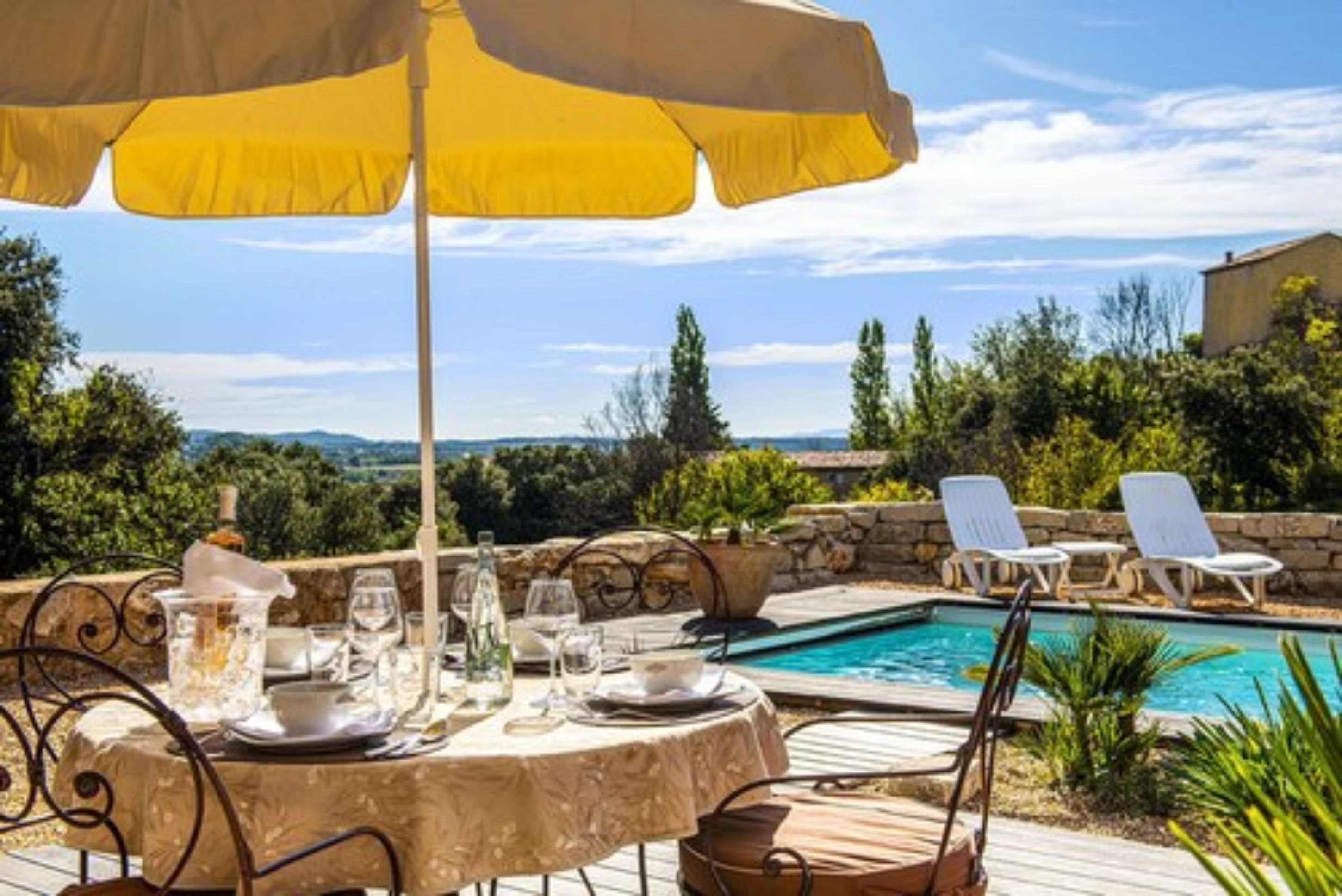 Un espace repas aménagé au bord de la piscine, avec une table dressée pour deux personnes, un parasol jaune et une vue sur la piscine et la végétation environnante.