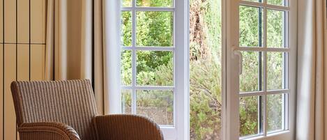 Une chaise en osier placée près d’une grande fenêtre donnant sur un paysage verdoyant.