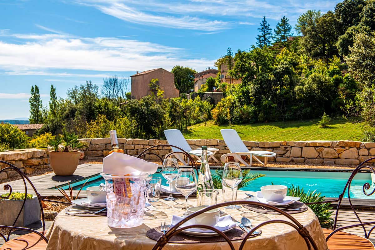 A poolside dining area with a table set for two, overlooking a swimming pool and lush greenery.