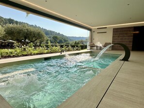 A modern indoor pool with a waterfall feature, surrounded by a landscaped garden and overlooking a scenic view of hills. - La Bastide du Calalou Hotel & Spa, BW Premier Collection (Moissac-Bellevue)