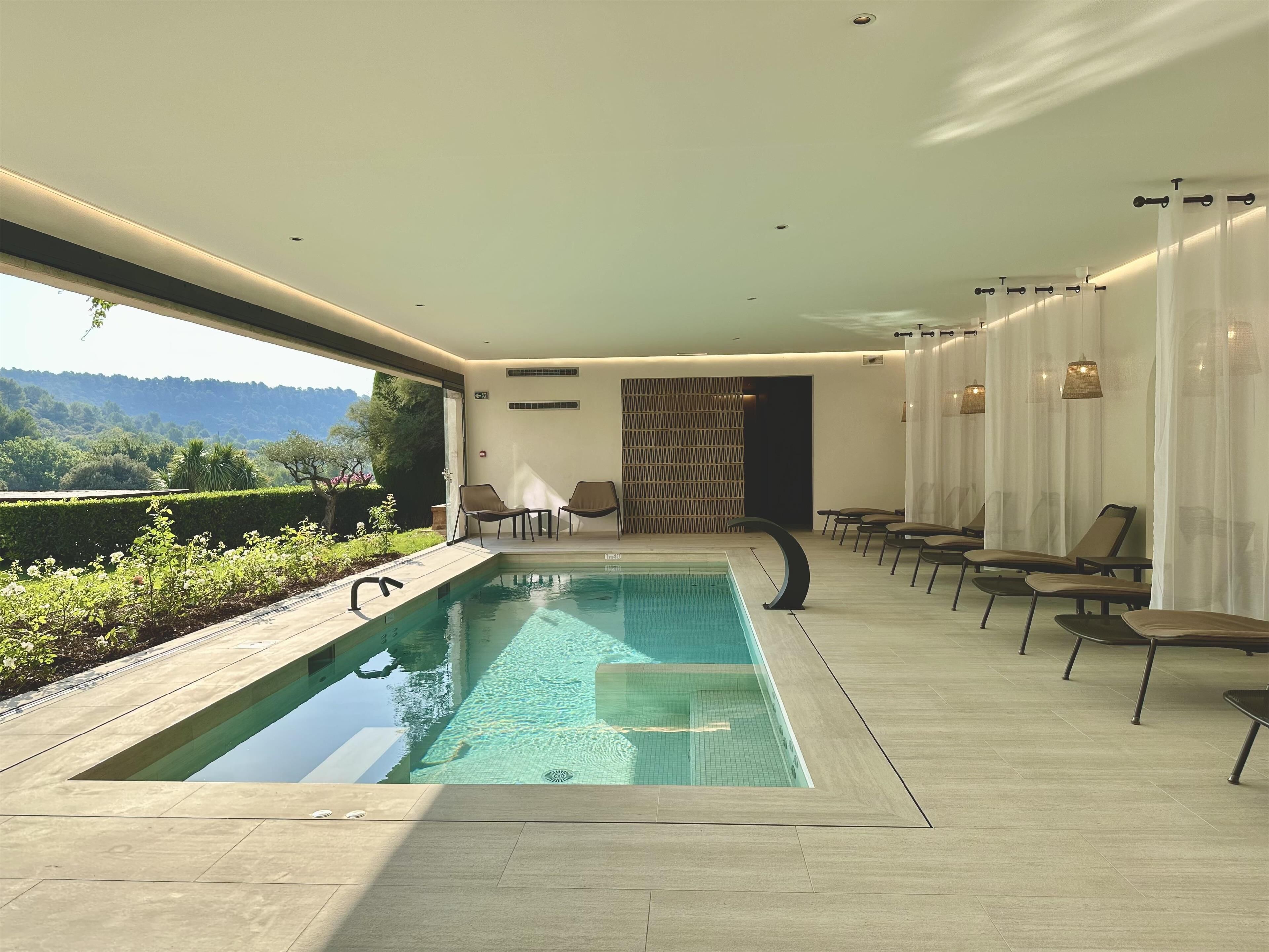 Un espace piscine intérieur moderne, doté de parois en verre transparent, d’une piscine et de chaises longues.