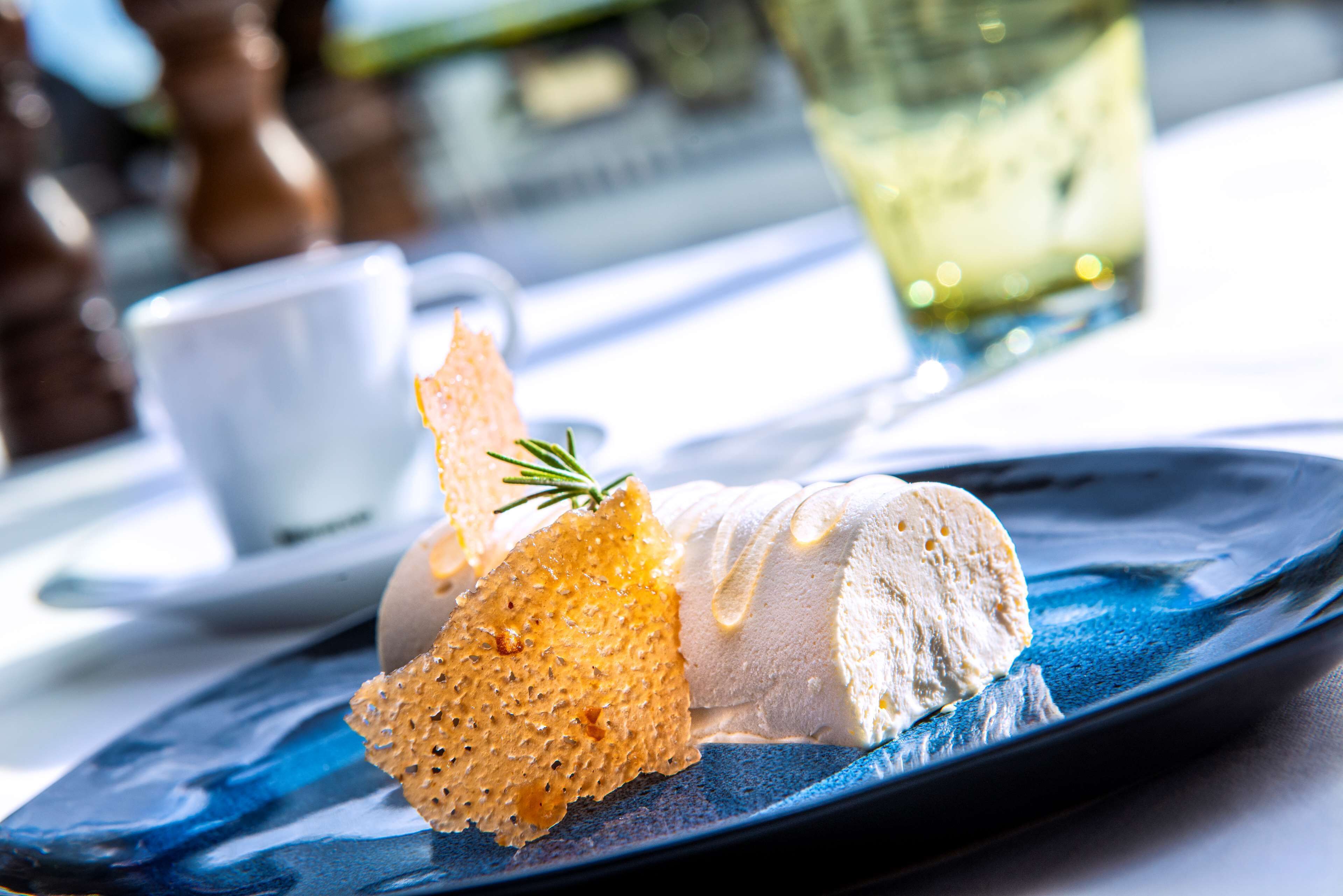 A dessert on a blue plate with a sprig of rosemary, a white cup, and a glass of a yellowish drink on a table.