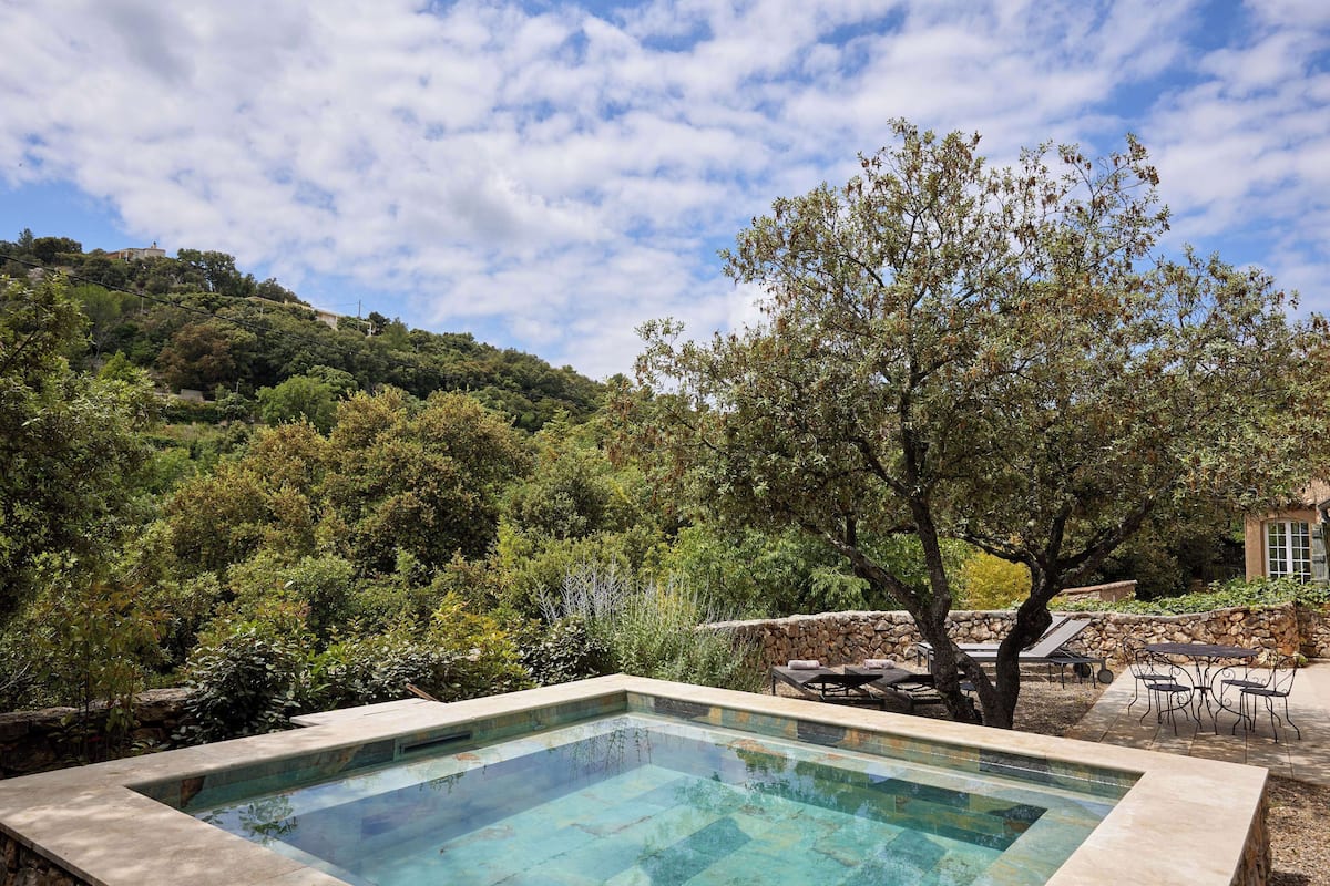 A rectangular pool with clear water, surrounded by a stone wall and a tree.
