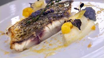 Une assiette blanche avec un morceau de poisson, des garnitures jaunes et violettes, et une branche d’herbe aromatique.