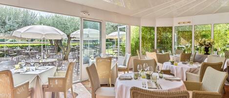 An outdoor dining area with wicker chairs and tables, large umbrellas, and a view of a garden.