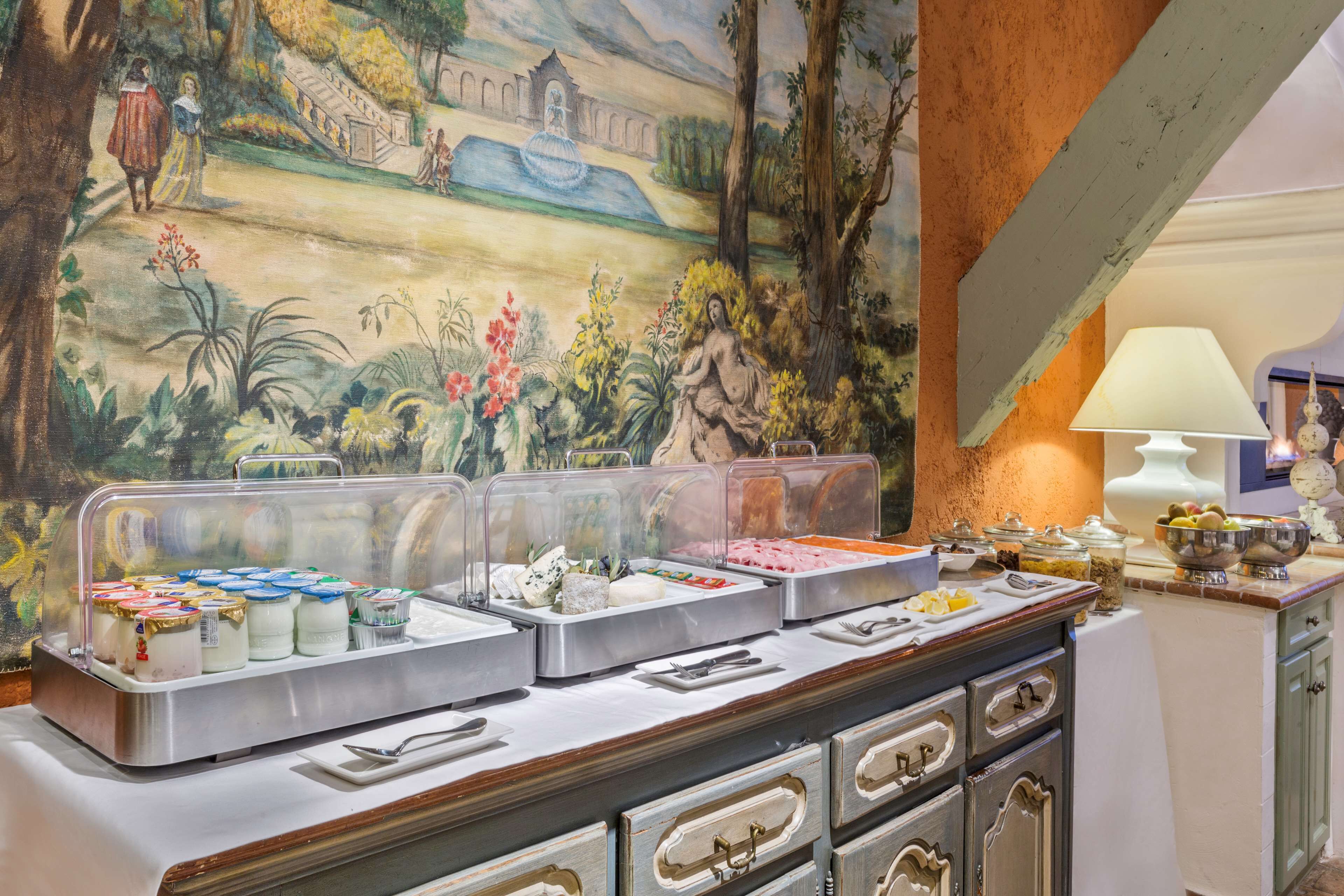 A buffet with various food items in clear containers, set against a mural of a landscape with figures and architecture.