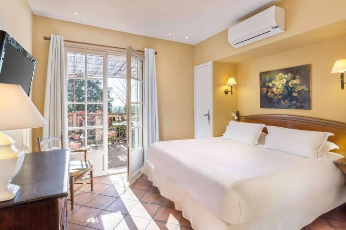 A hotel room with a bed, a desk, a chair, a window with curtains, an air conditioning unit, and a painting on the wall.