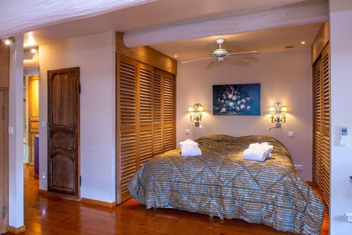 A bedroom with a bed, wooden shutters, a ceiling fan, wall-mounted lights, and a painting.