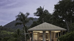 A two-story wooden structure with a balcony, surrounded by lush greenery and palm trees, overlooking a hilly landscape. - Kapuhala (Koh Samui)