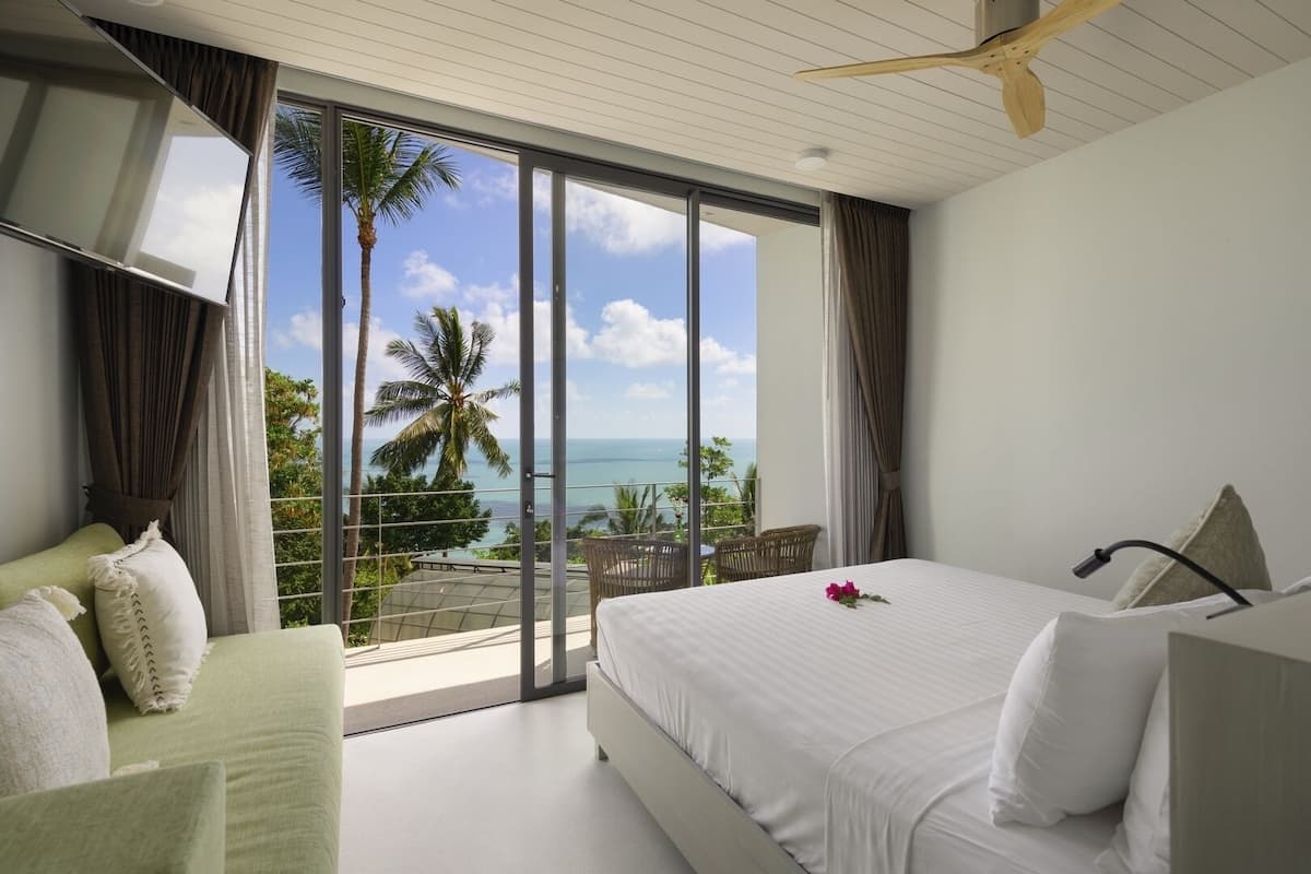 Une chambre d’hôtel avec un grand lit, un ventilateur de plafond et une vue sur l’océan et les palmiers.
