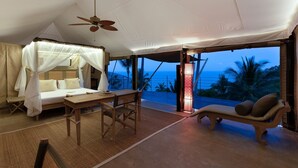 A tented room with a bed, a table, and a chair, overlooking a beach at dusk. - Kapuhala (Koh Samui)