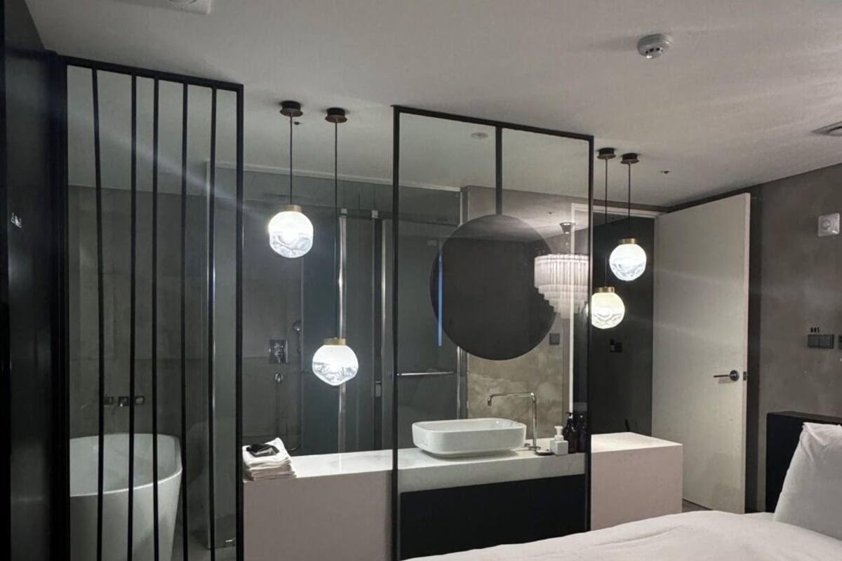 A modern hotel room with a large bed, a bathroom with a bathtub, and a mirror with three pendant lights.
